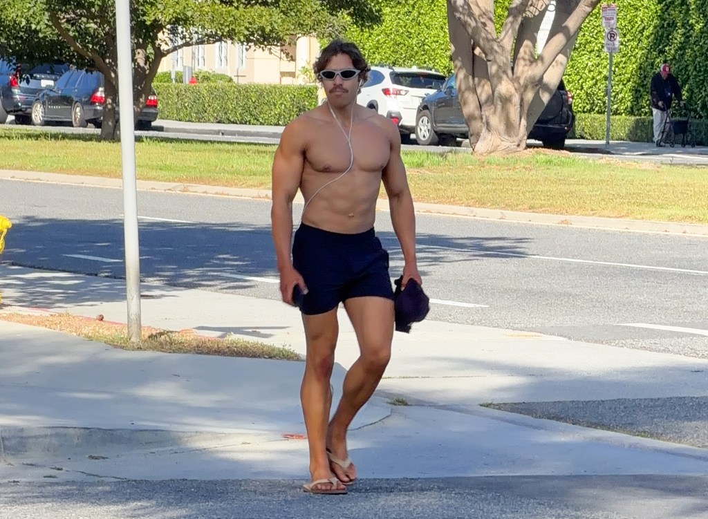 Joseph Baena walks shirtless in shorts, sunglasses, and earbuds.