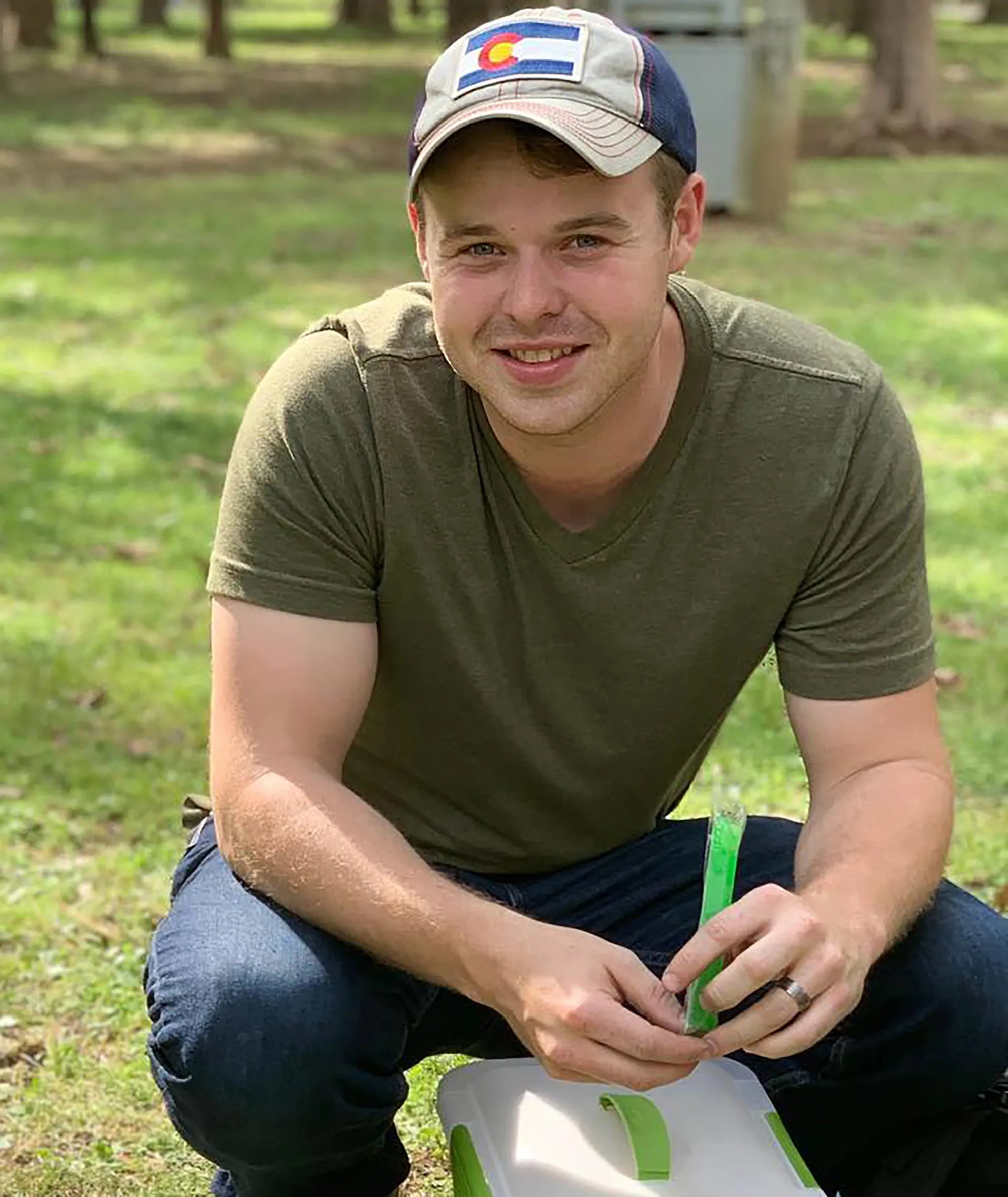 Joseph Duggar in a gray and blue Colorado hat, green shirt, and jeans, smiling while crouching outdoors.