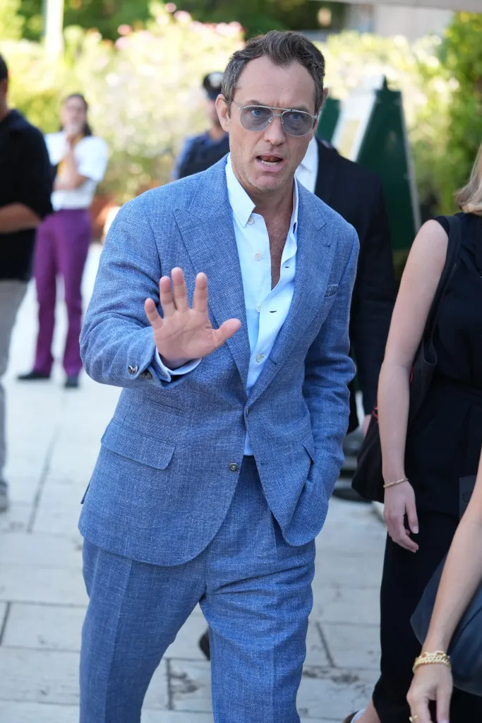 Jude Law in a blue suit and sunglasses, holding up his hand at the 82nd Venice International Film Festival.
