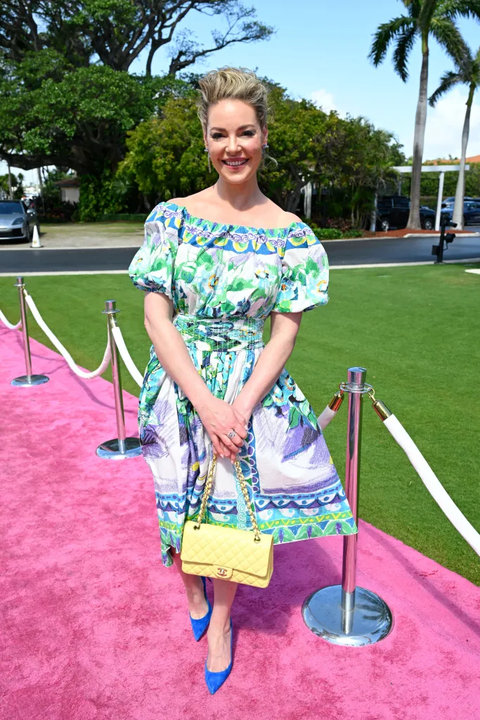 Katherine Heigl attends the Wine, Women & Shoes Event at Mar-a-Lago, wearing a colorful off-the-shoulder dress, holding a yellow quilted handbag, and standing on a pink carpet.