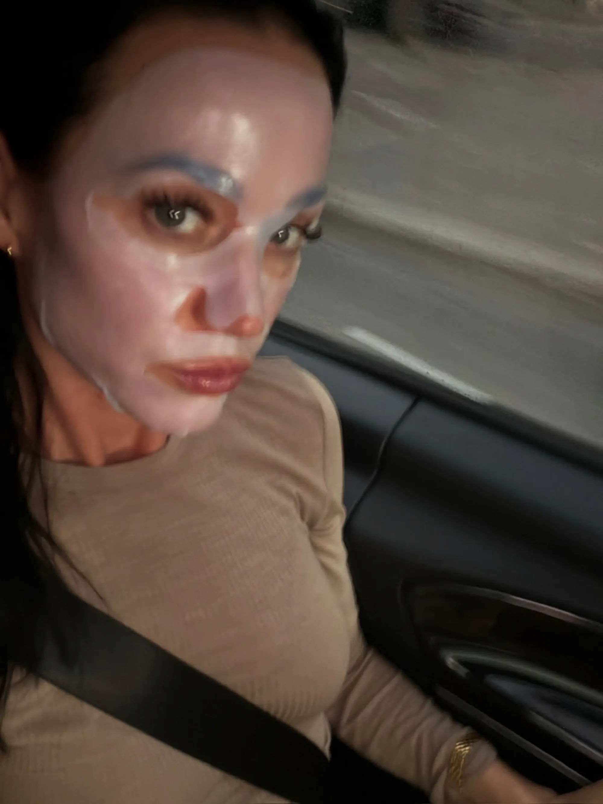A woman wearing a sheet mask and seatbelt in a car.