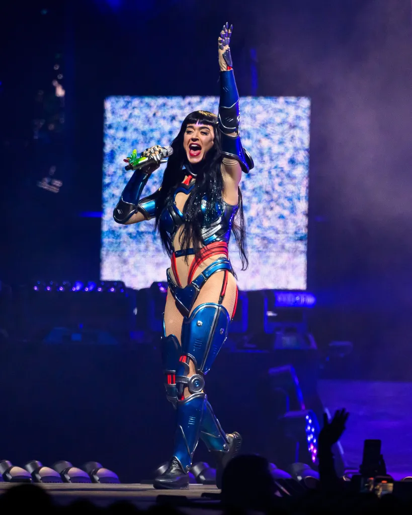 Katy Perry performing live in a futuristic blue and red armored outfit with a robotic arm, holding a colorful microphone.