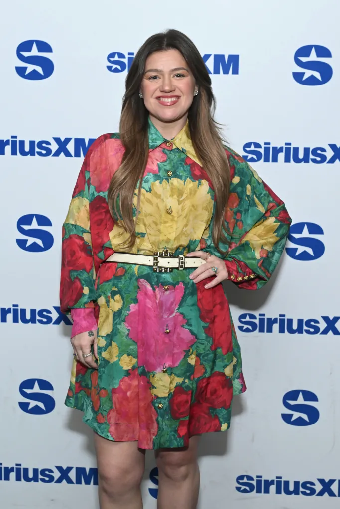 Kelly Clarkson smiling in a vibrant floral dress with a white belt, standing in front of a white background with blue SiriusXM logos.