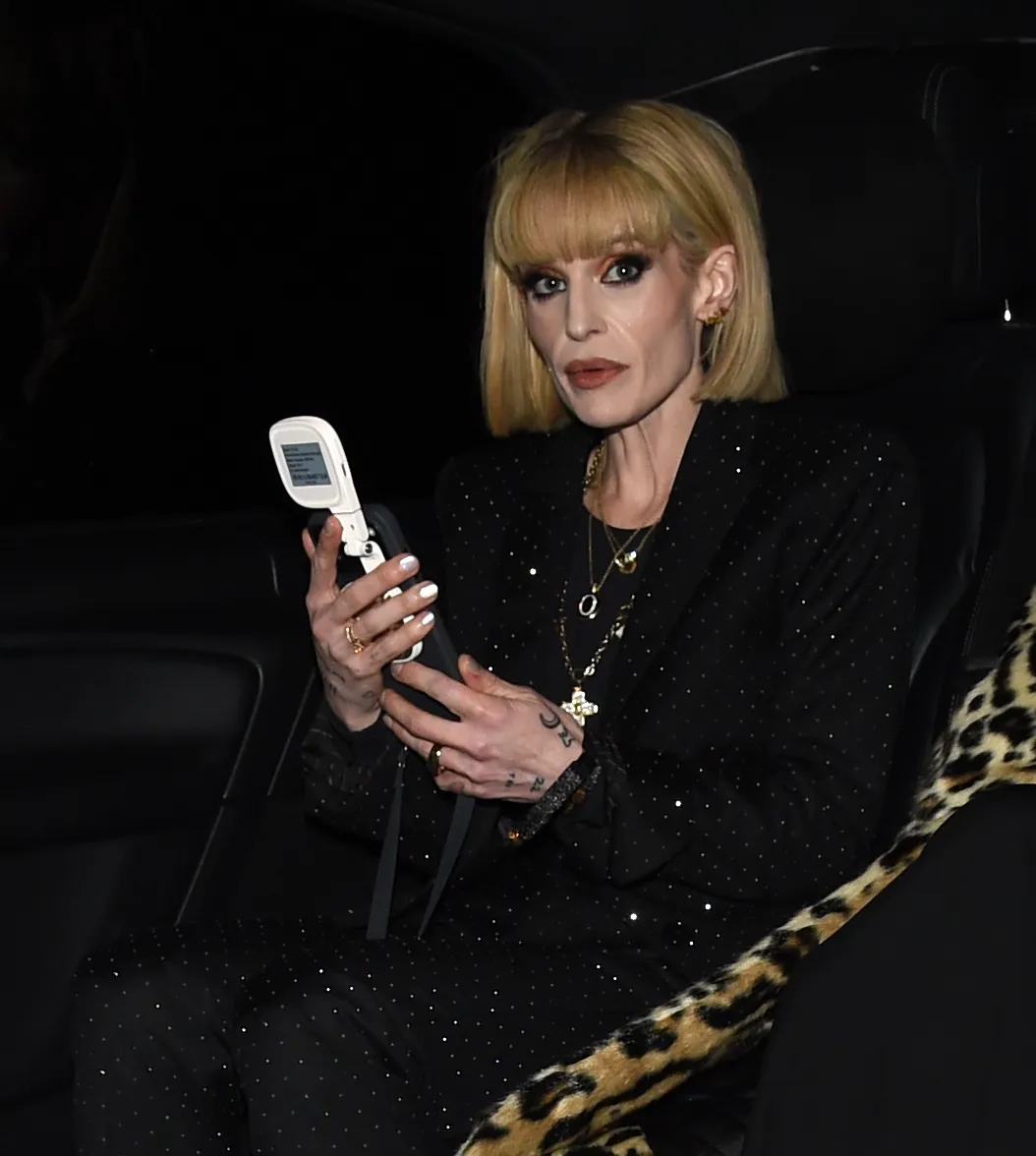 Kelly Osbourne in a sparkling black suit holding a phone, with a leopard print item beside her.