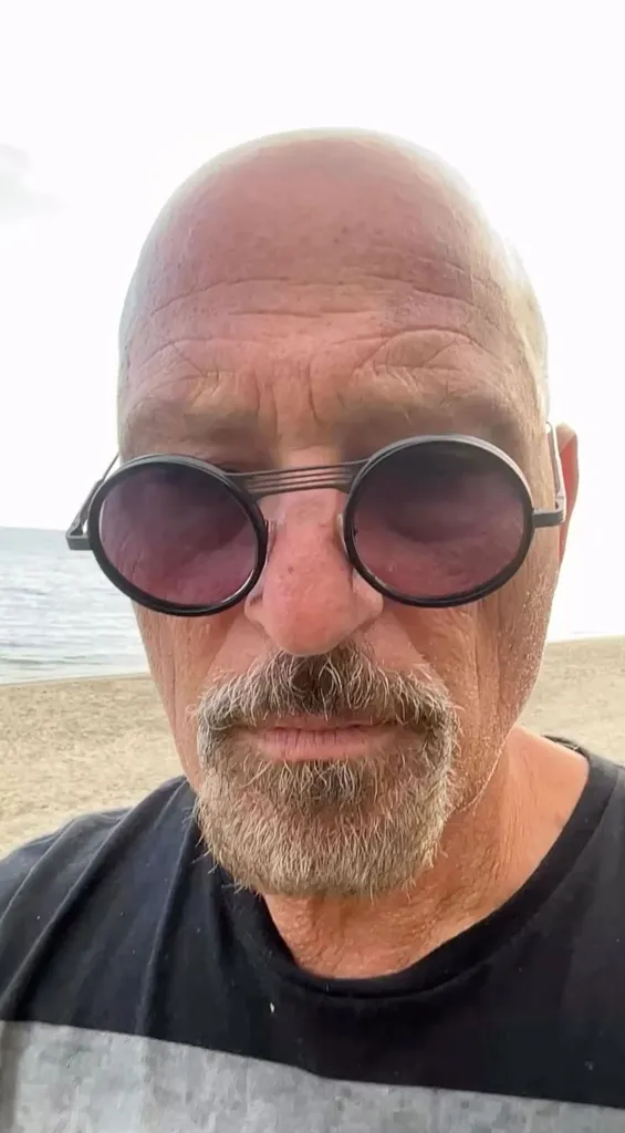 Howie Mandel in sunglasses and a black t-shirt, taking a selfie at a beach.