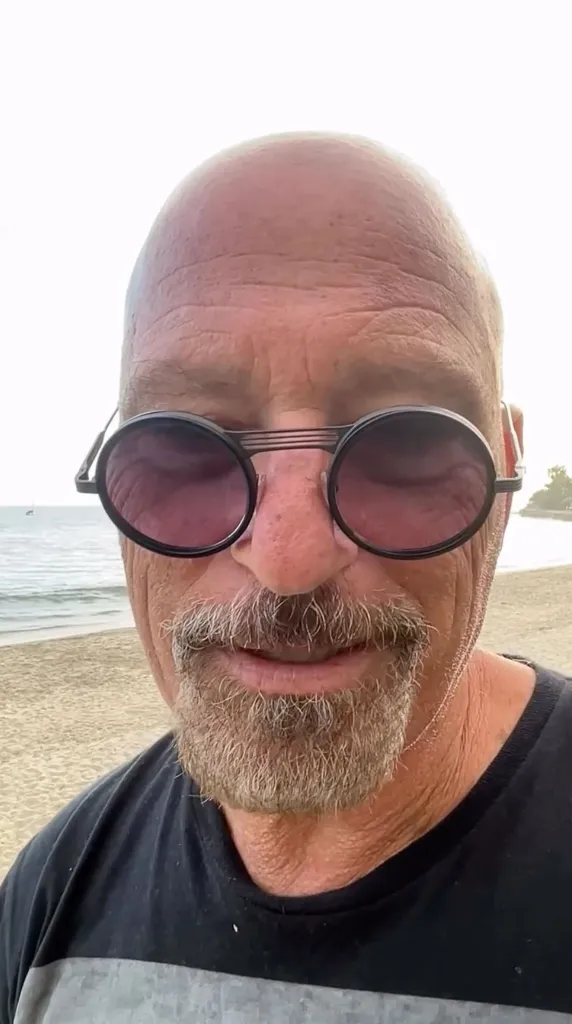 Howie Mandel wearing round sunglasses on a beach.