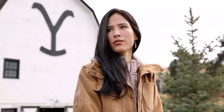 Kelsey Asbille as Monica Dutton from Yellowstone, standing outside in a brown jacket.