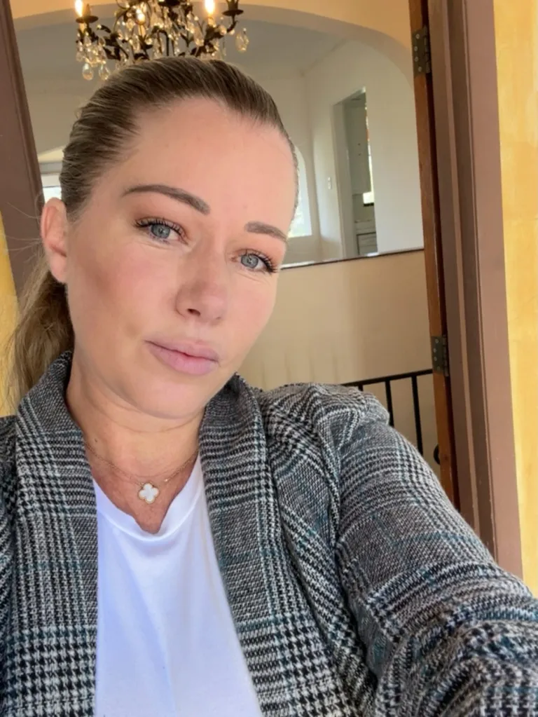Kendra Wilkinson looking at the camera wearing a gray plaid blazer over a white top and a clover pendant necklace.