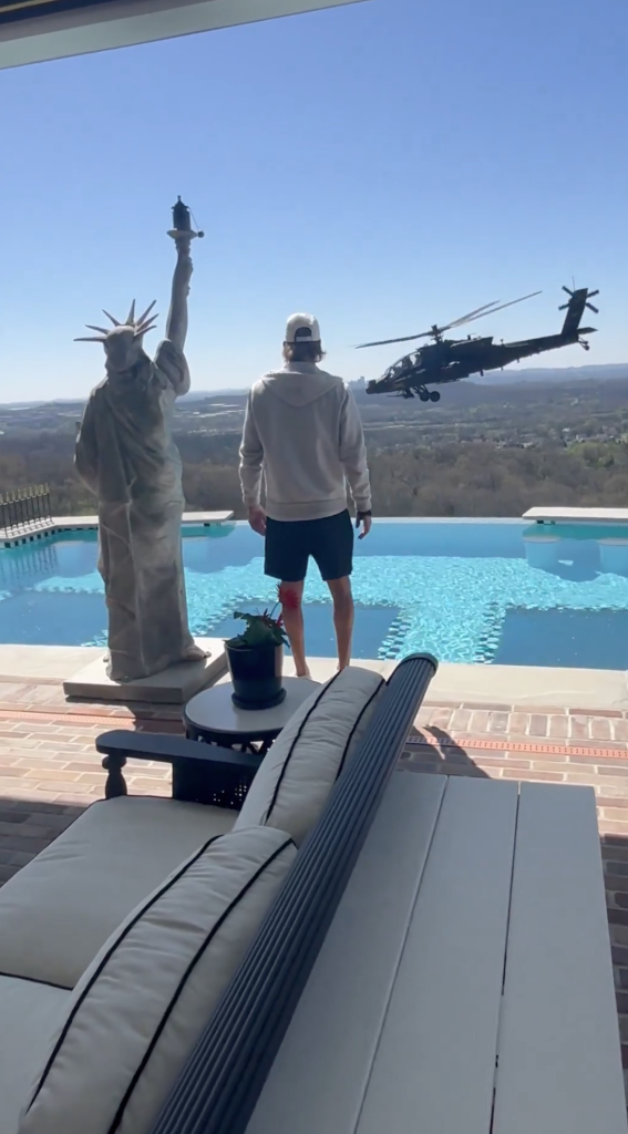 Kid Rock looking at a military helicopter flying over his pool with a Statue of Liberty replica.