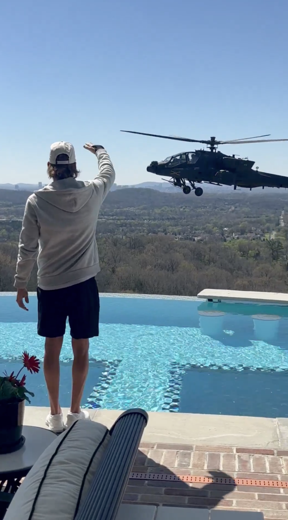 Kid Rock waving at a military helicopter flying over an infinity pool overlooking a valley.