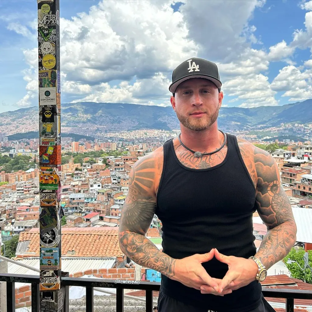 Chet Hanks wearing a black tank top and baseball cap, with tattoos on both arms, poses in front of a city landscape.