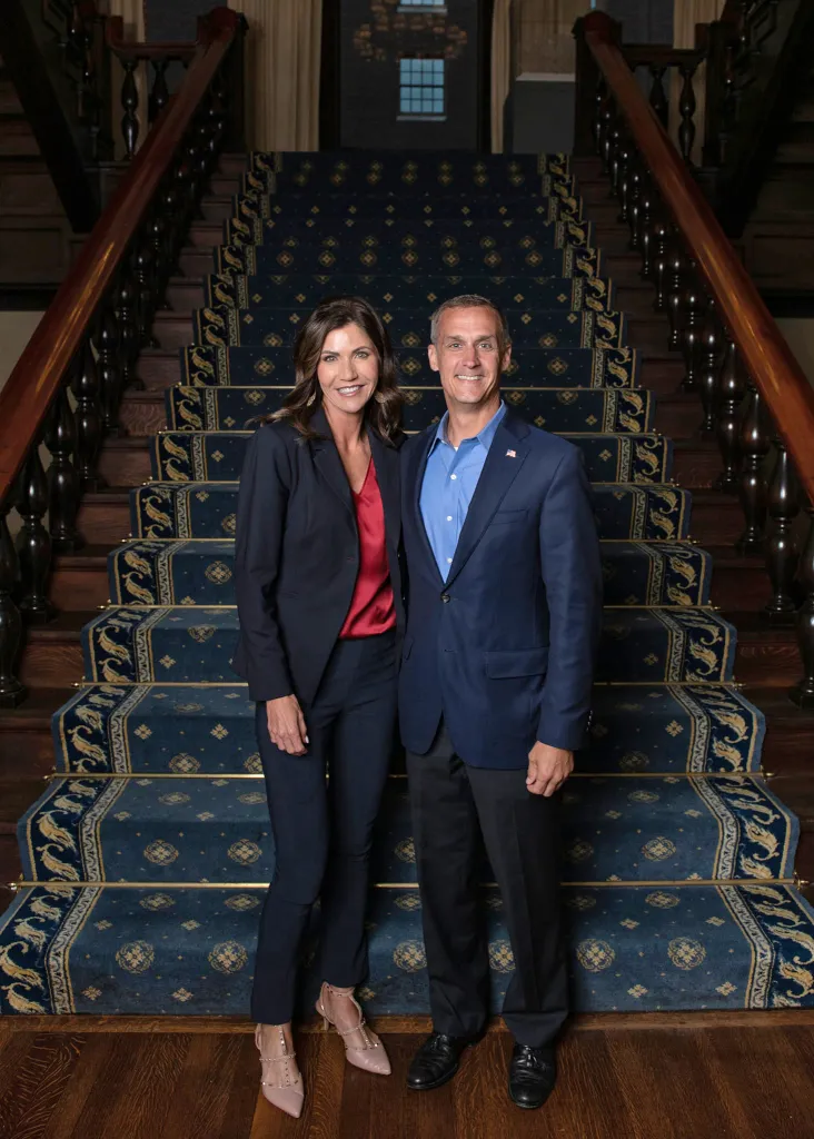 Kristi Noem and Corey Lewandowski smiling while standing at the bottom of a grand staircase.