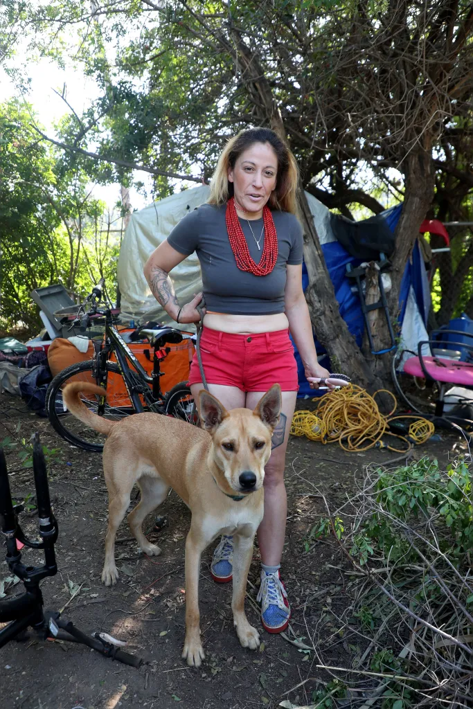 Kween, a woman with tattoos, stands with her dog in front of her makeshift shelter by the Los Angeles River.