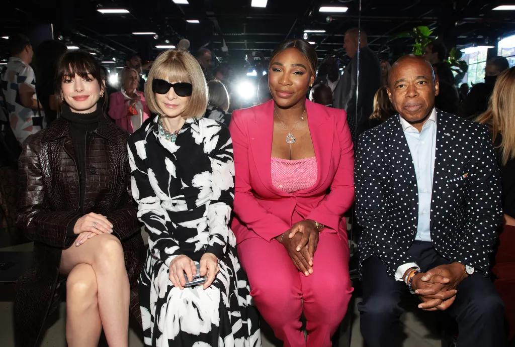 Anne Hathaway, in a black dress and leather jacket, Anna Wintour, in a white and black patterned dress and sunglasses, Serena Williams, in a pink pant suit and pink top, and Eric Adams, in a black and white polka dot jacket, blue shirt and black pants, sit in a front row.