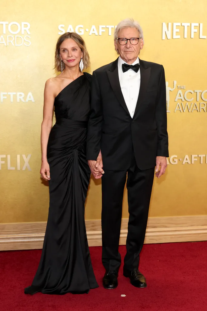 Calista Flockhart in a black one-shoulder gown and Harrison Ford in a black tuxedo hold hands on a red carpet.