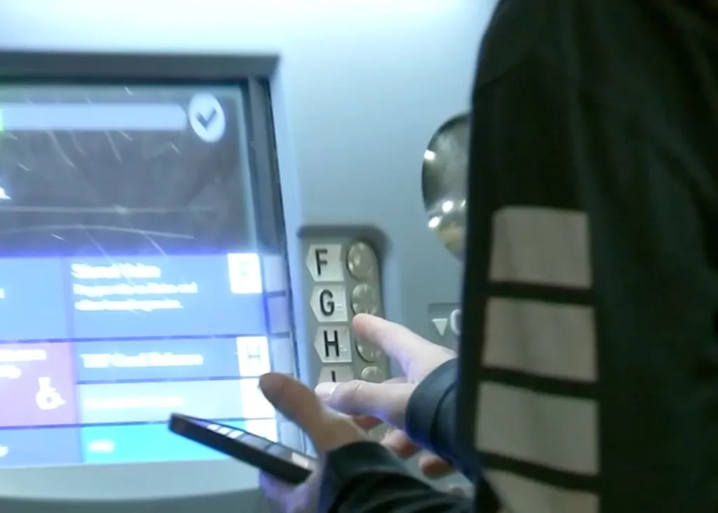 A person selects an option on a touchscreen ticket machine while holding a smartphone.