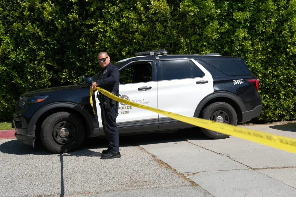 A police officer with sunglasses stands by a police SUV, holding yellow police tape to block off a road.