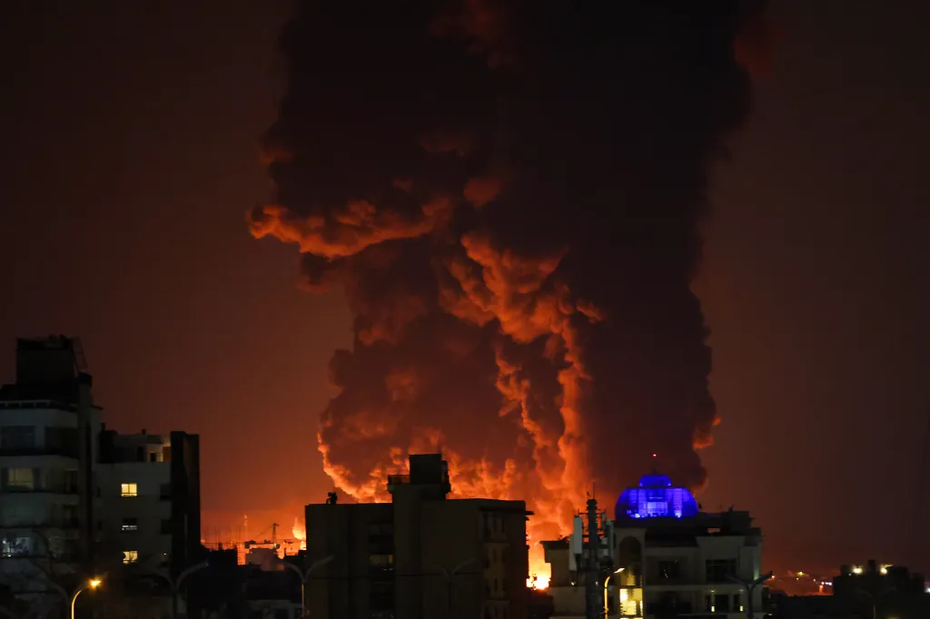 Large plumes of smoke and fire rise above buildings in Tehran at night.