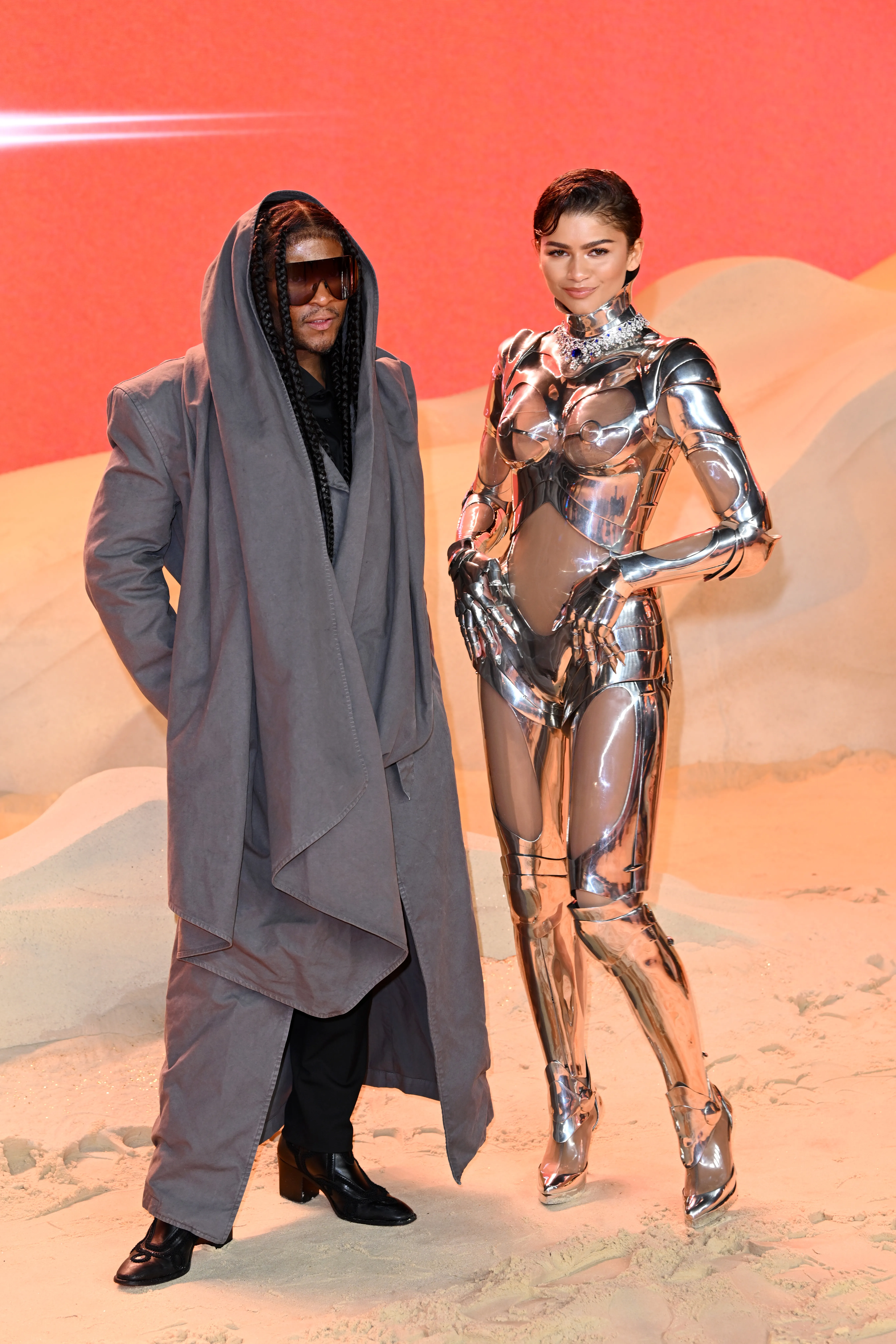 Law Roach wearing a gray robe and Zendaya in a metallic robot-like suit at the 