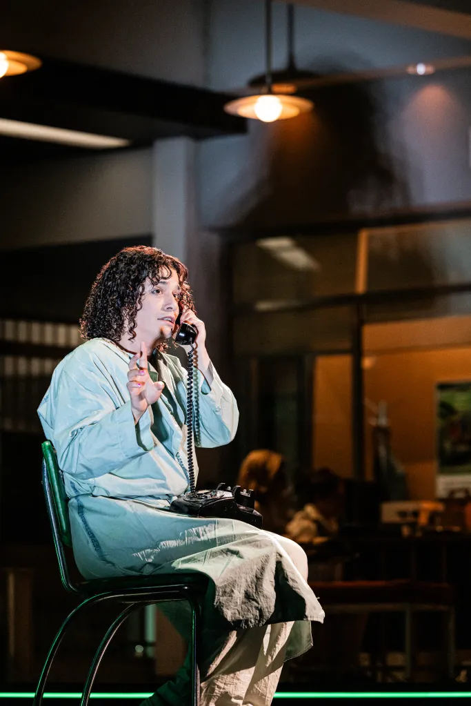 Esteban Andres Cruz as Leon sitting on a green chair, wearing a light blue smock and holding a rotary phone to his ear.