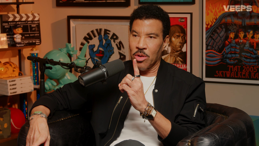 Lionel Richie sitting in a chair in front of a microphone with posters in the background.