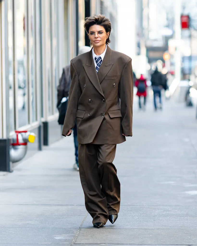 Lisa Rinna wearing a brown pantsuit with a striped tie and glasses, walking on a city sidewalk.