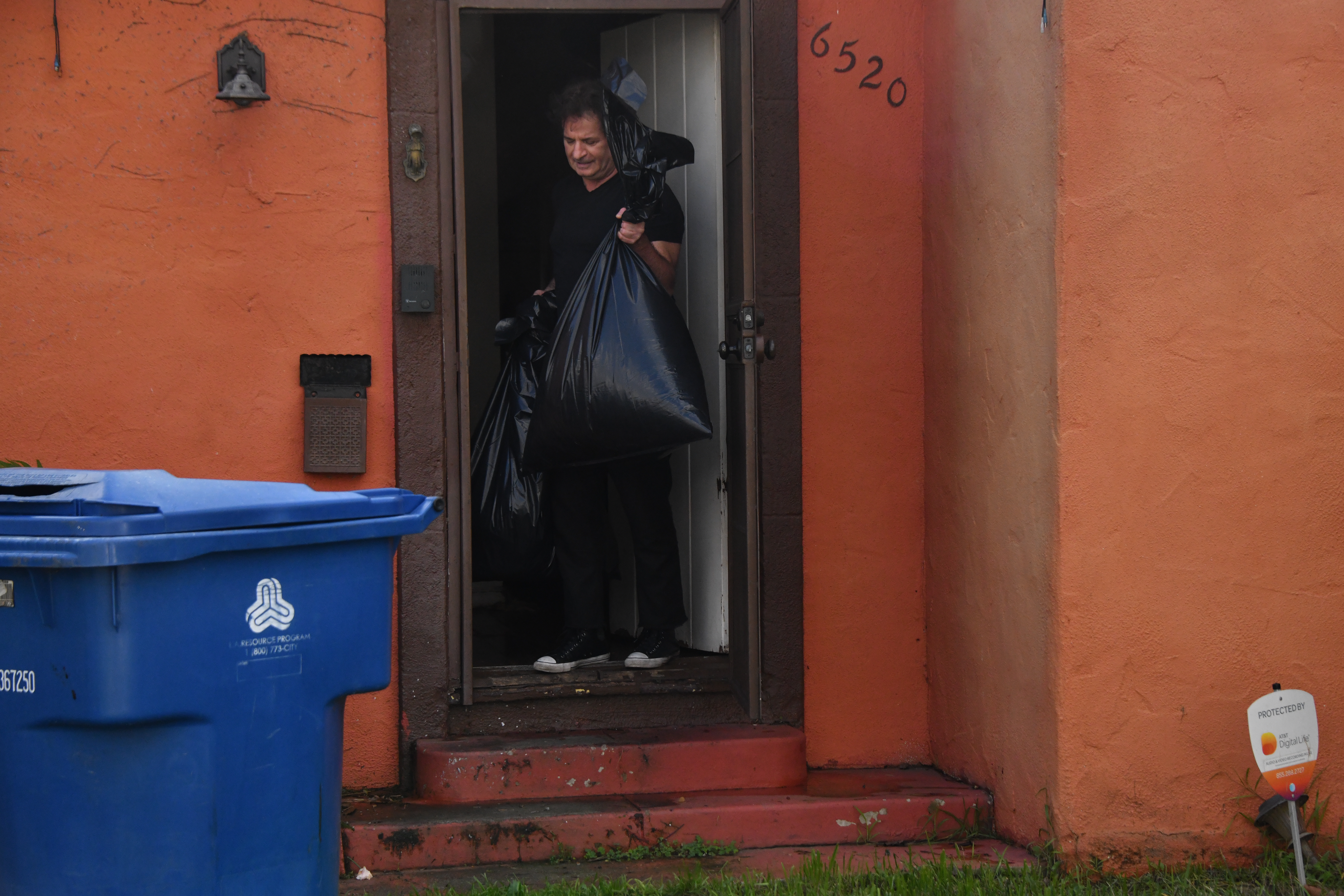 A person carries two large black garbage bags out of a doorway next to a blue recycling bin.