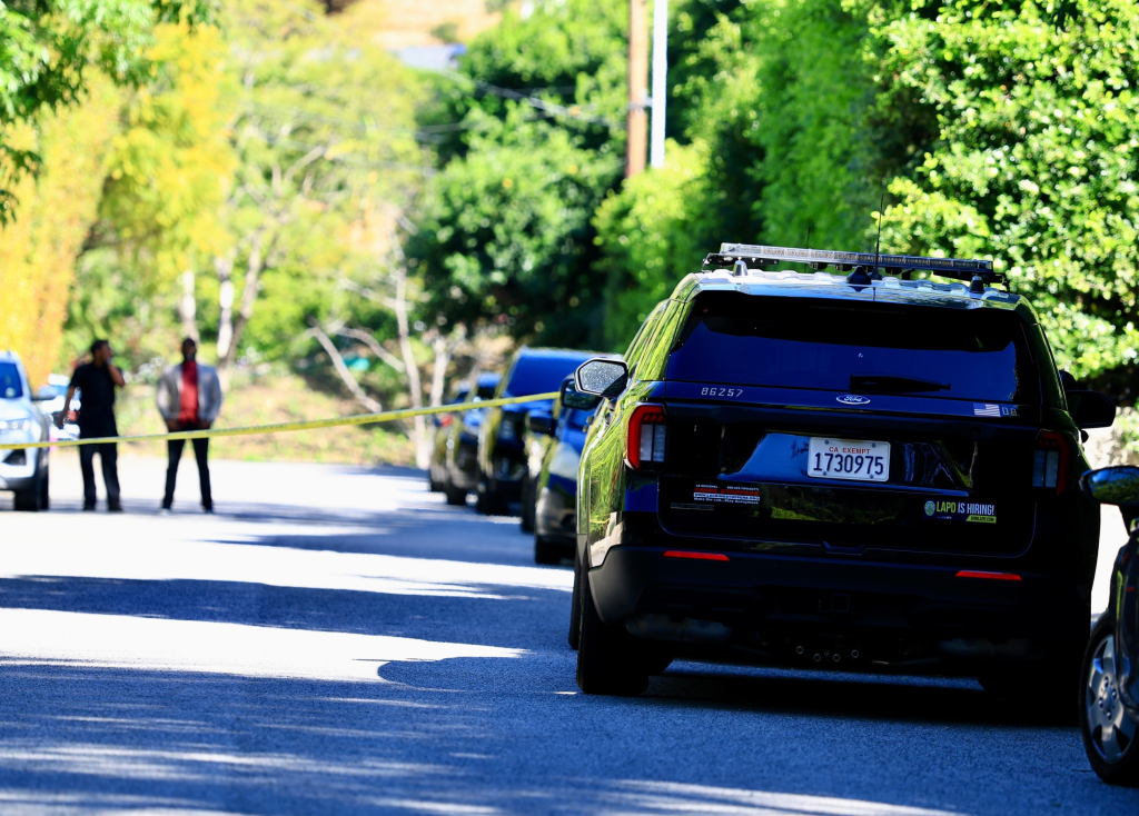 General view of a street with a police vehicle, several cars, and two people behind a yellow tape, possibly indicating an incident or investigation.