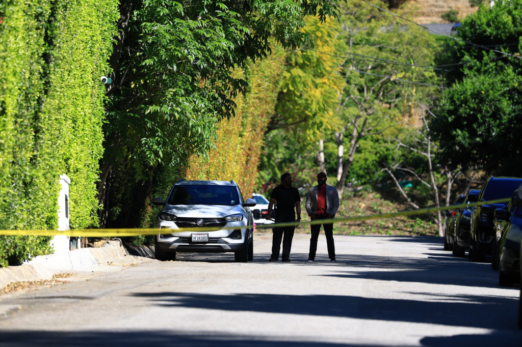 A white SUV parked on a residential street blocked by yellow police tape, with two men standing behind it.