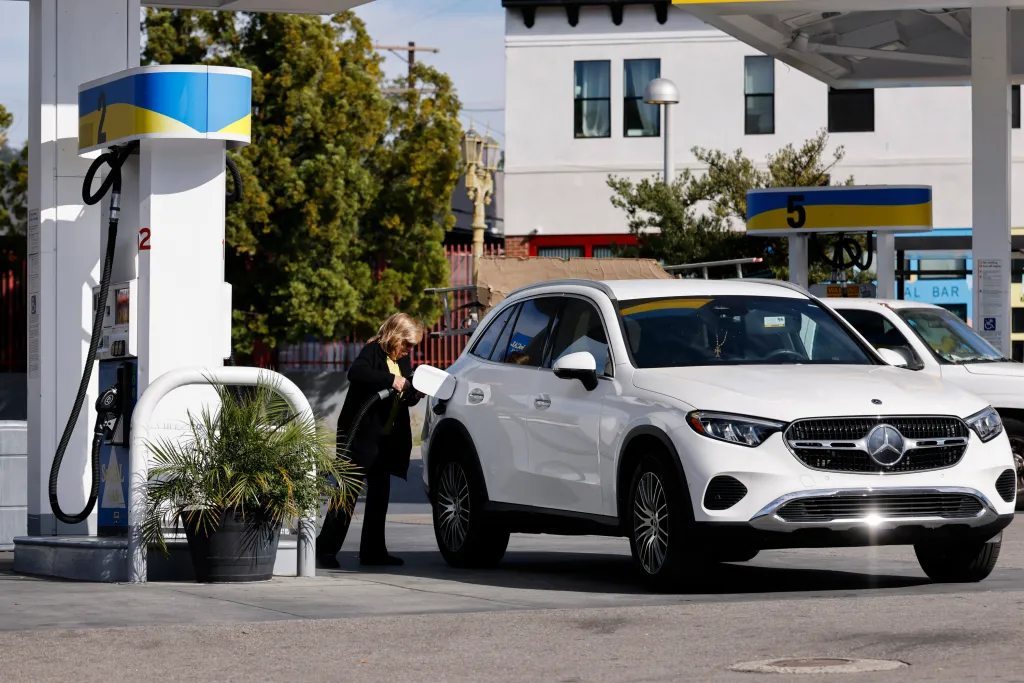A woman in a black jacket and yellow shirt pumps gas into a white Mercedes-Benz SUV at a SoCal Gas station.