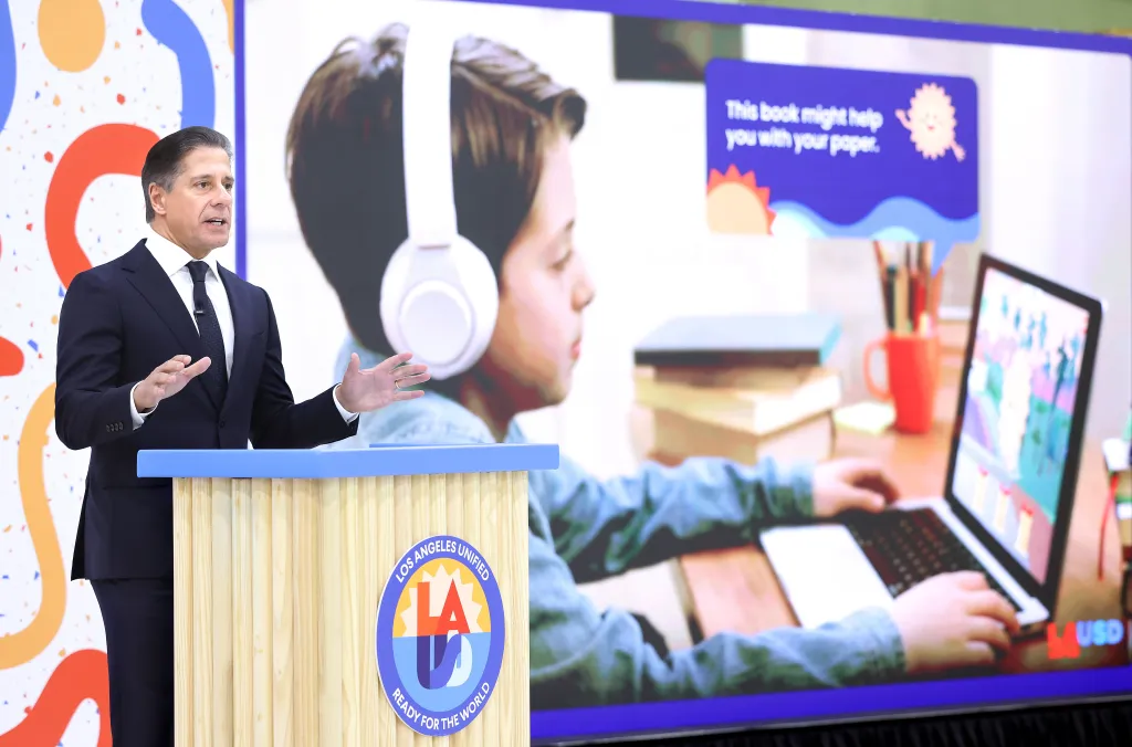 Los Angeles Unified Supt. Alberto Carvalho speaks at a podium with a screen showing a child using an AI-assisted learning platform in the background.