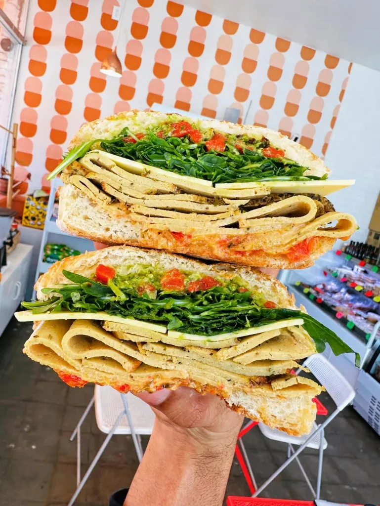 A hand holding up a large plant-based sandwich, cut in half, filled with arugula, plant-based cheese, and plant-based deli slices.
