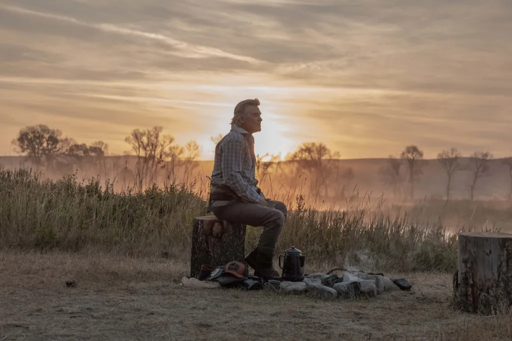 Kurt Russell sitting on a tree stump by a campfire at sunrise.