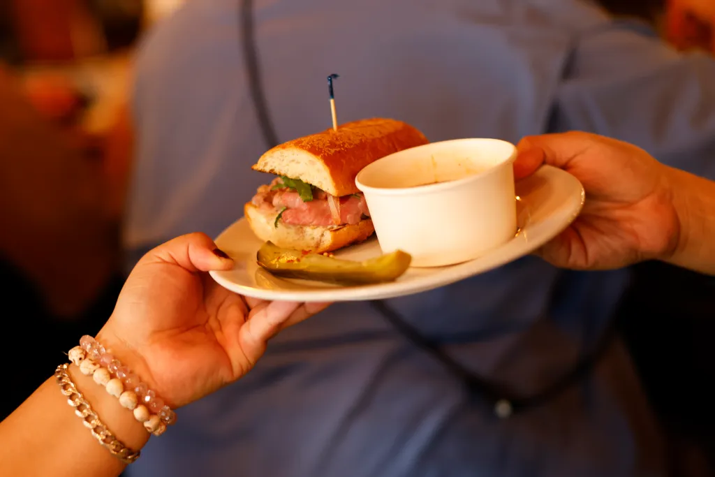 Two hands holding a plate with a French dip sandwich, a pickle spear, and a small cup of dipping sauce.