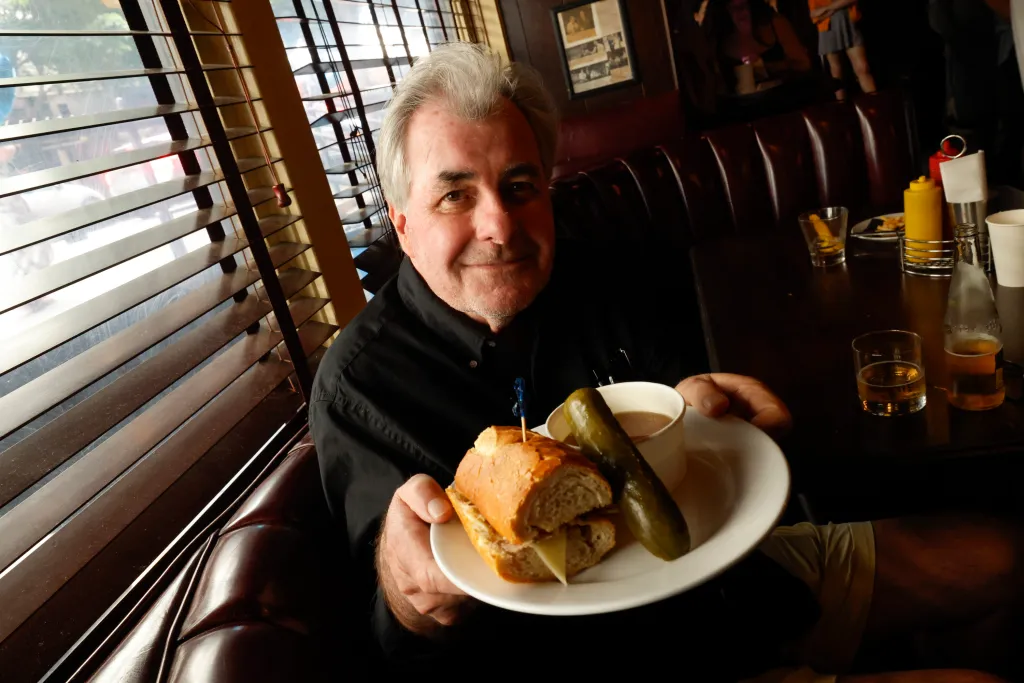 Thomas Haggerty holding a braised pork French dip, pickle, and cup of au jus at Cole's French Dip.