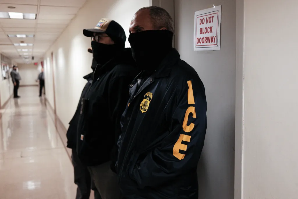 Two masked federal agents wearing ICE jackets stand in a hallway.