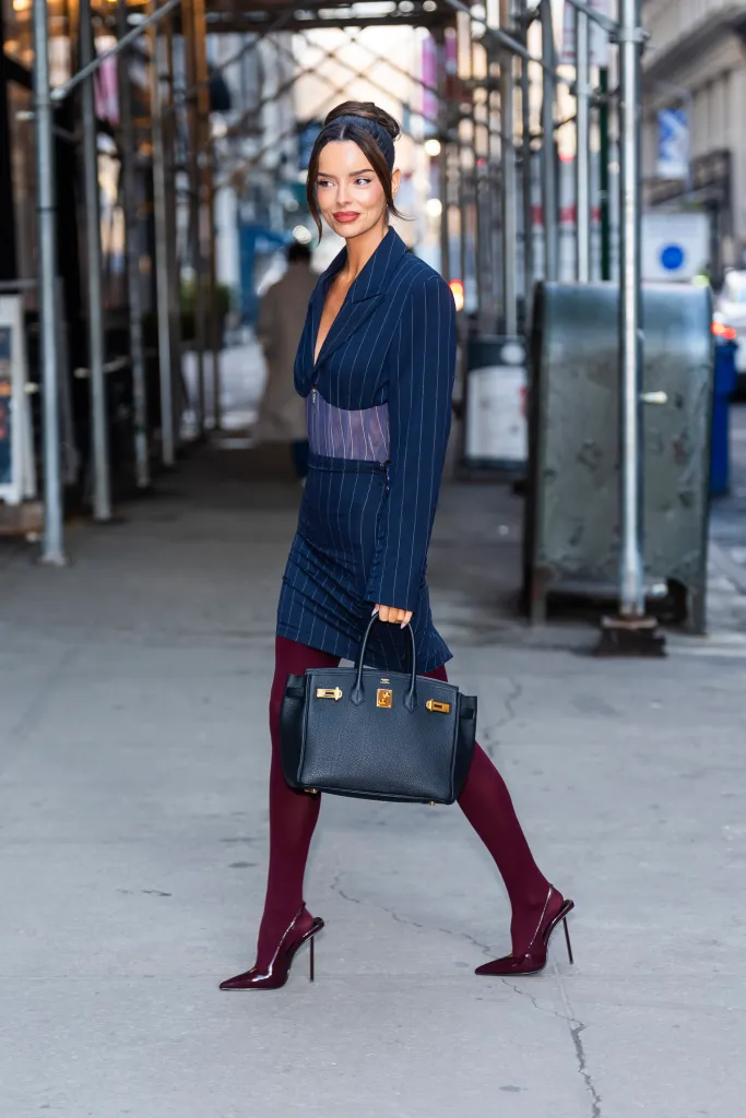 Maura Higgins in a pinstripe blazer dress, maroon tights and heels, and a black handbag in New York City.