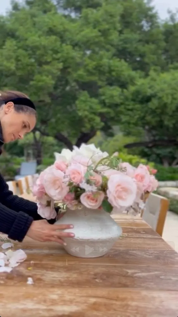 Meghan Markle arranging pink flowers in a vase on a wooden table.