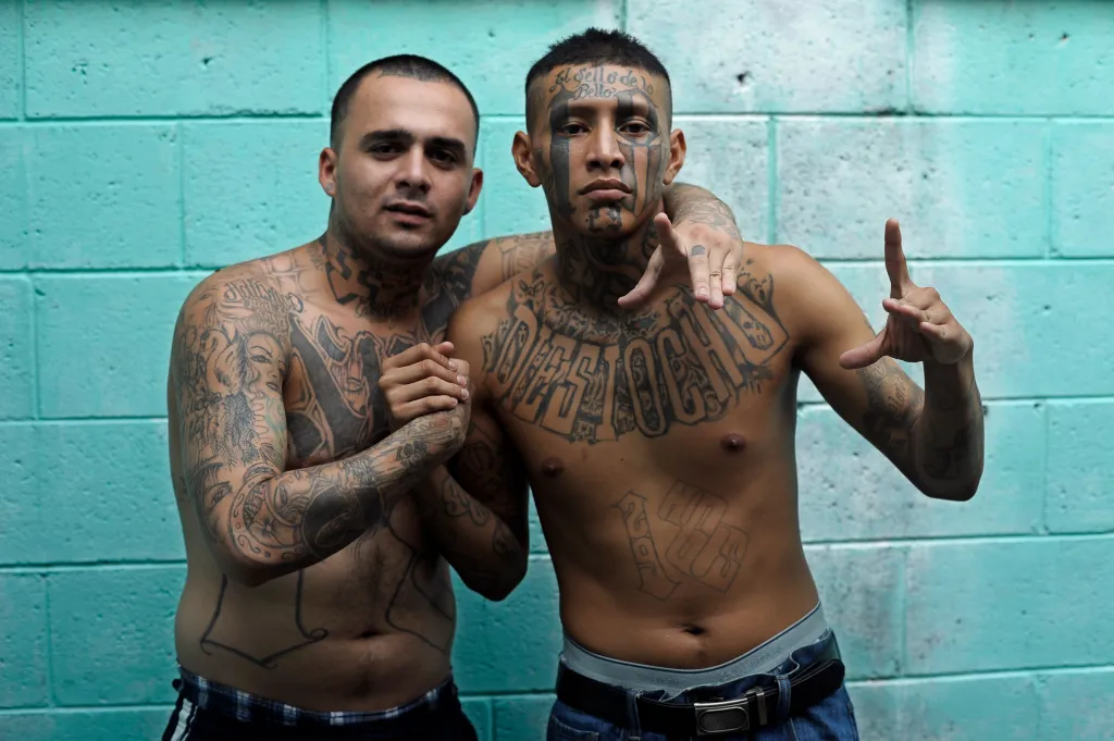 Two shirtless men covered in tattoos, one pointing, against a turquoise wall.