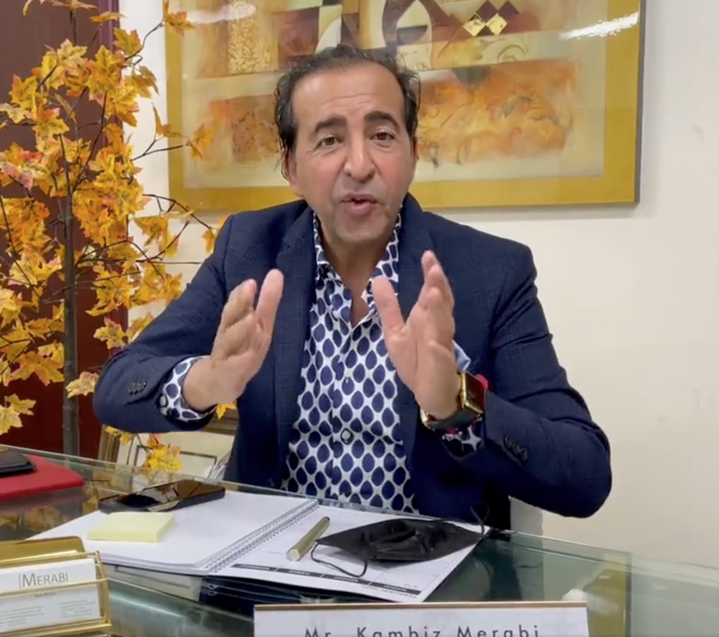 A man in a suit gesturing with his hands while sitting at a desk with his name, Mr. Kambiz Merabi, on a sign.