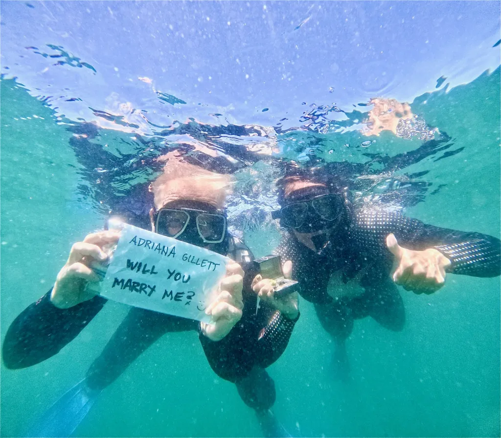 An underwater photo of James Hetfield proposing to Adriana Gillett with a handwritten sign and ring box.