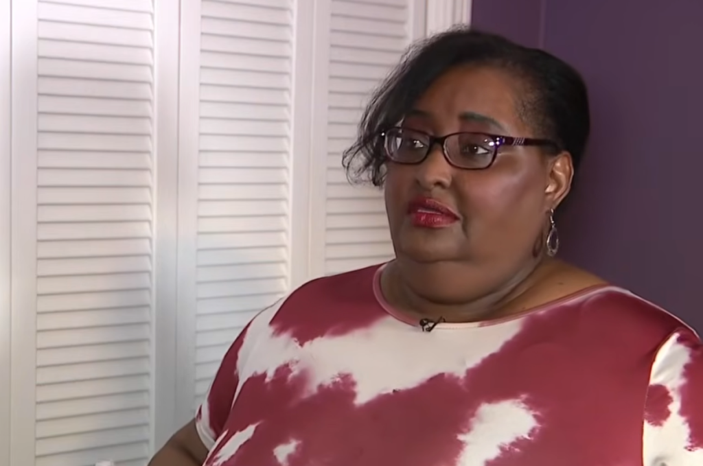 A dark-skinned woman with glasses and short, dark, curly hair wearing a white and red patterned shirt.