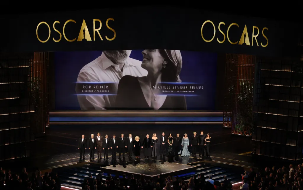 Celebrities on stage at the Oscars in front of a screen displaying an image of Rob Reiner and Michele Singer Reiner.