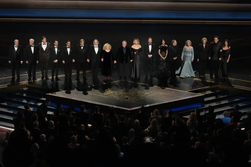 Seventeen actors stand on a dark stage, some holding hands, at the Dolby Theatre.