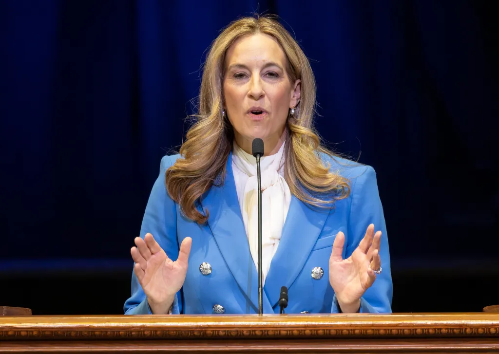 Mikkie Sherrill speaking at her inauguration, dressed in a blue blazer and white blouse, with a microphone in front of her.