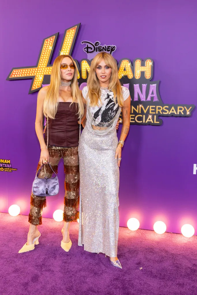 Alex Cooper and Miley Cyrus pose on a purple carpet.