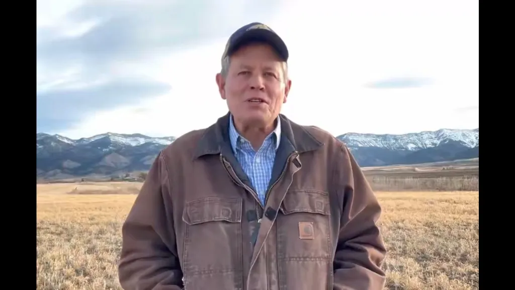 Steve Daines standing outdoors in a brown jacket and blue cap with mountains in the background.