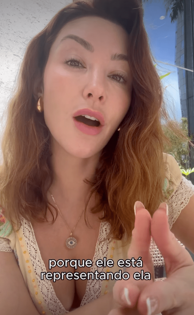 A woman with brown hair, wearing a white shirt and a necklace, speaks while making a gesture with her hand.