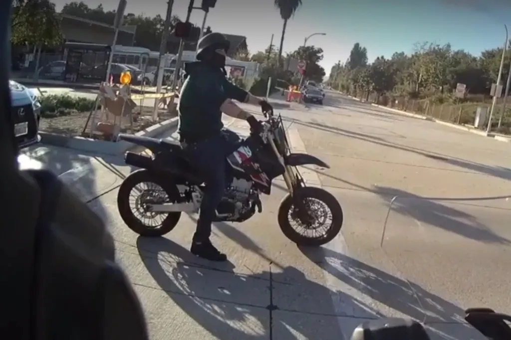A motorcyclist wearing a helmet and dark clothing sits on a dirt bike at an intersection, with buildings and palm trees in the background.