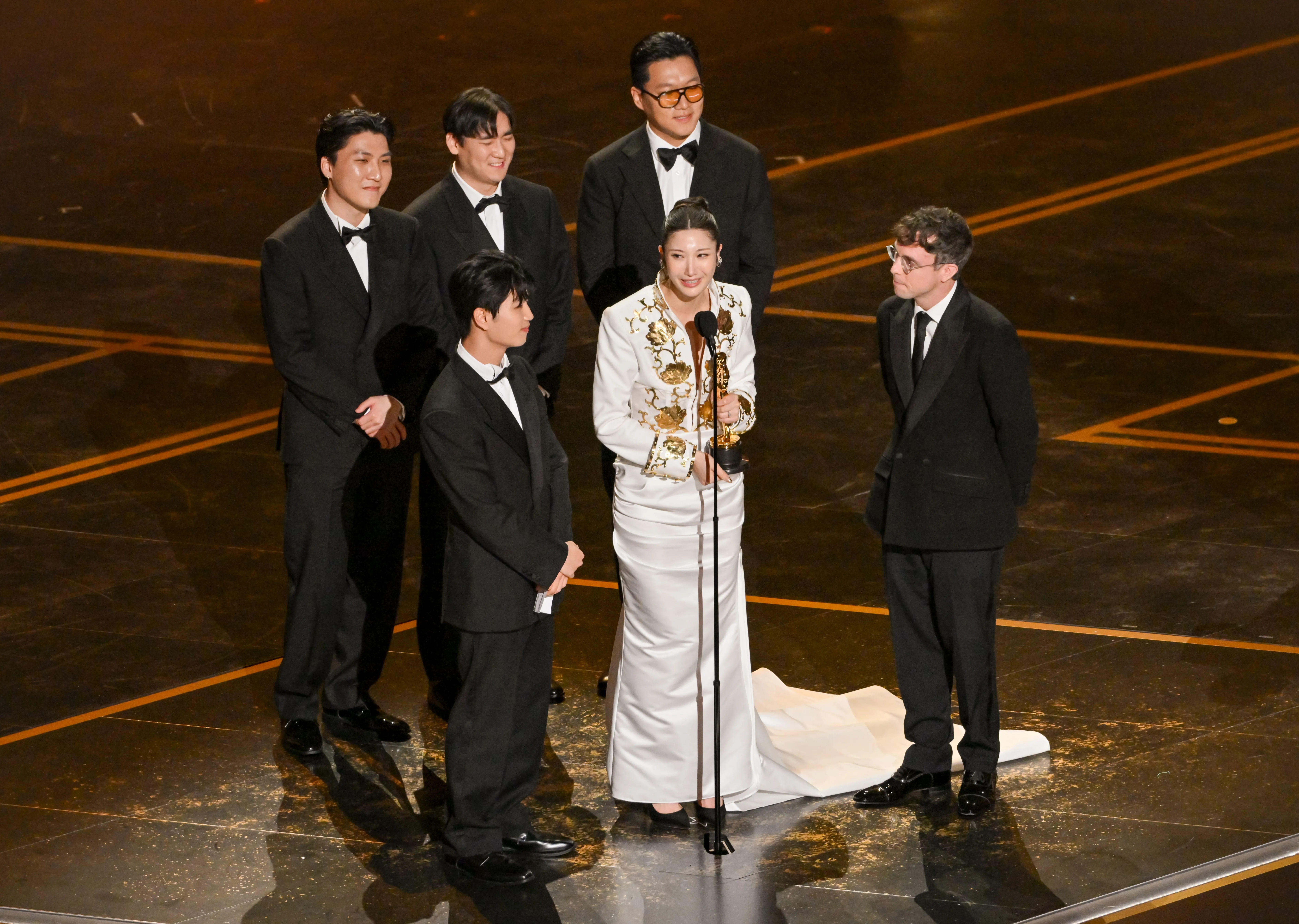 A woman in a white gown and gold top holds an Oscar statue while speaking into a microphone, surrounded by five men in tuxedos